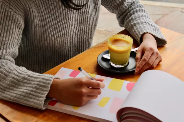 Bilden visar en person som sitter vid ett bord med en kopp kaffe. Framför personen finns ett anteckningsblock.